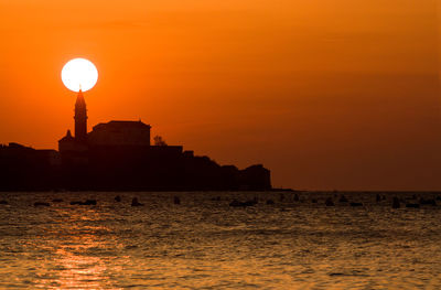 Silhouette buildings by sea against orange sky