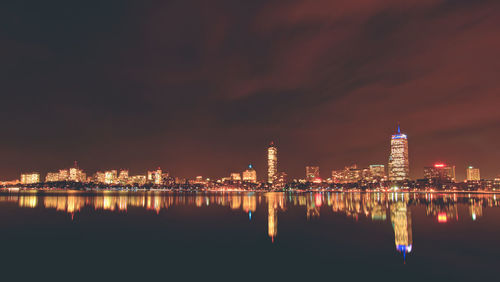 Illuminated buildings by river against sky at night