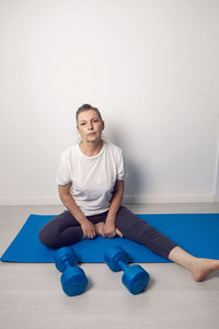 Elderly female athlete is sitting at home on a yoga mat and with dumbbells