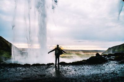 Rear view of silhouette man standing on rock against waterfall