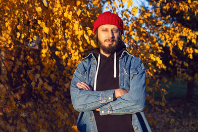 Hipster man with a beard in jeans and a knitted red hat is stand on a woodpile in autumn