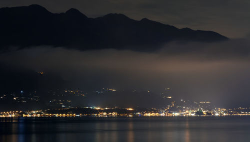 Illuminated city by sea against sky at night
