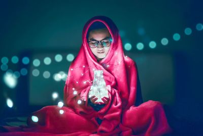 Young woman wrapped in blanket holding jar with illuminated string light at home