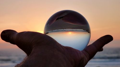 Close-up of hand holding crystal ball against sky during sunset