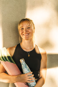 Happy woman holding water bottle and exercise mat against wall