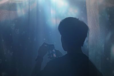 Rear view of man photographing through mobile phone amidst curtains