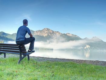 Man sit on wooden bench at coast of picturesque lake bellow blue mountain summits. calmness