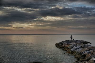 Distant view of man standing on rocks by sea against cloudy sky during sunset