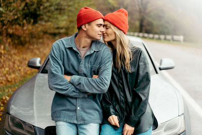 A young man and woman embrace tenderly while sitting on the bonnet of a car.