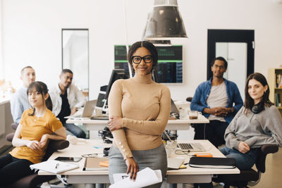 Portrait of smiling businesswoman standing with confident colleagues sitting at desk in creative office