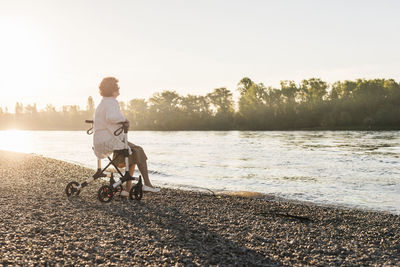 Pensive senior woman sitting on her wheeled walker at riverside