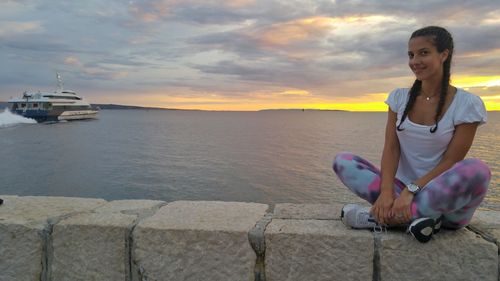 Portrait of young woman sitting on shore against sky during sunset
