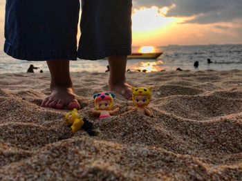 Low section of kid standing by toys on sand at beach against sky during sunset
