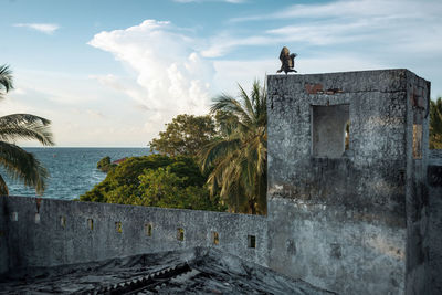 Ruin of villa by sea against sky with bird taking off