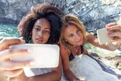 Smiling two women taking selfie while lying on rock formation at sea