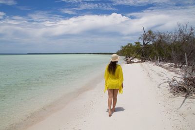 Rear view of young woman walking at beach against cloudy sky