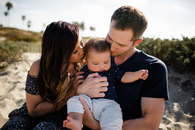 Mother & father kissing infant son while sitting on beach