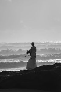 Bride with bouquet standing on shore against sky