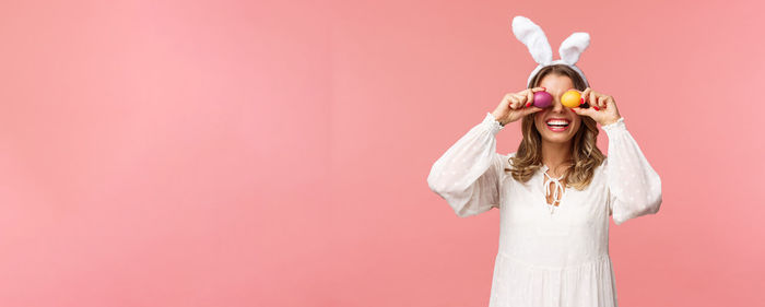 Portrait of woman covering eyes with eggs against coral background