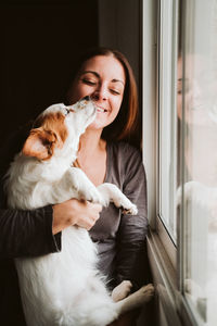 Smiling woman with dog standing by window at home