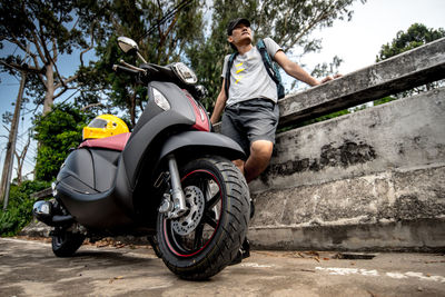 Low angle view of man standing by retaining wall and motor scooter