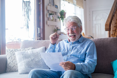 Cheerful senior man holding paper while sitting on sofa at home