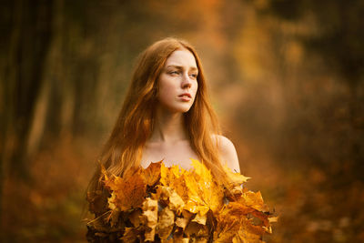 Thoughtful young woman covered with leaves standing at forest during autumn