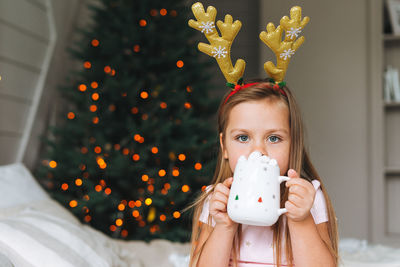 Cute smiling funny little girl in pink dress drinking cocoa sitting on bed in room with christmas tr