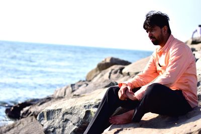 Man sitting on rock by sea against clear sky