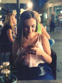 Smiling young woman using smart phone while sitting at sidewalk cafe at night