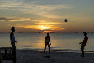 Men playing with ball on beach against sky during sunset