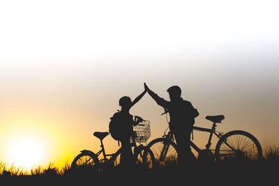 Silhouette friends with bicycles giving high-five while standing on grassy field against clear sky during sunset