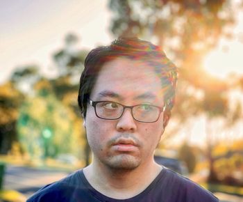 Portrait of young asian man in eyeglasses looking away against setting sun and trees.