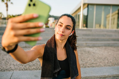 Young slim female in sportswear with earbuds and fitness tracker taking selfie on mobile phone while standing on street after training