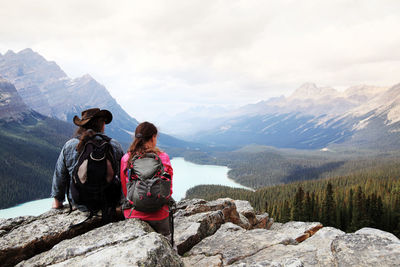 Rear view of man and woman sitting on rock formation overlooking mountains against sky