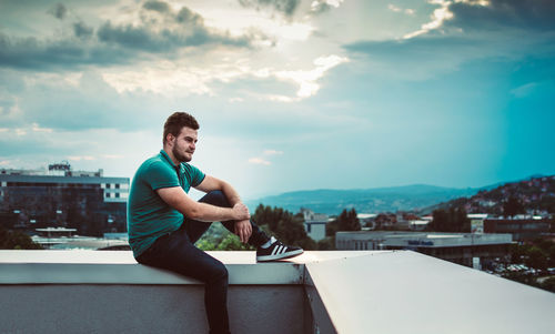 Young man sitting in city against sky