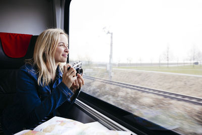 Happy blond woman with camera travelling by train looking out of window