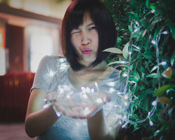 Portrait of young woman making face while holding illuminated lighting equipment by plant