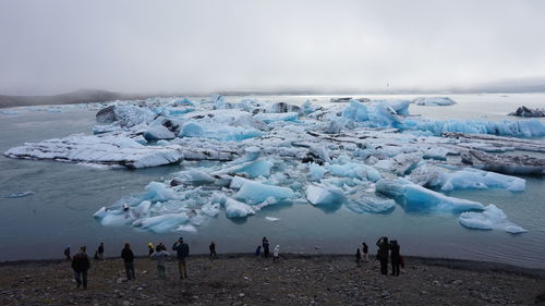 Tourists standing at beach by glaciers against sky