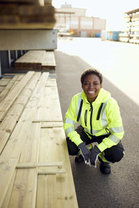 Portrait of smiling young female worker crouching by planks at lumber industry