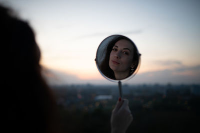 Close-up portrait of woman holding camera against sky during sunset