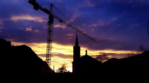 Silhouette buildings and crane against cloudy sky at dusk