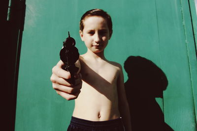 Portrait of shirtless boy holding toy gun against green wall during sunny day