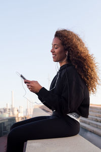 Side view of content african american female athlete in earphones text messaging on cellphone while having break from workout in city