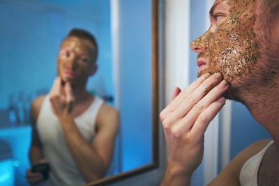 Close-up of man applying mask on face while looking in mirror at home