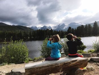 Rear view of children sitting by lake against sky