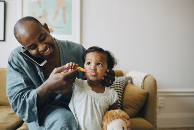 Man talking on mobile phone while brushing daughter's teeth at home