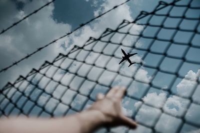 Cropped hand holding fence against airplane flying in sky