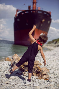 Fashionable boy child with long hair stand on a log next to a large ship that ran