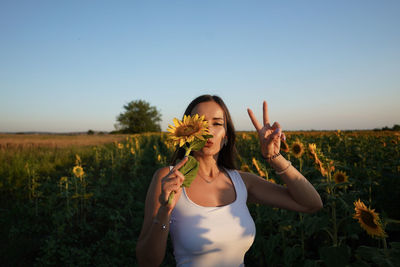Portrait of young woman holding sunflower gesturing against sky during sunset